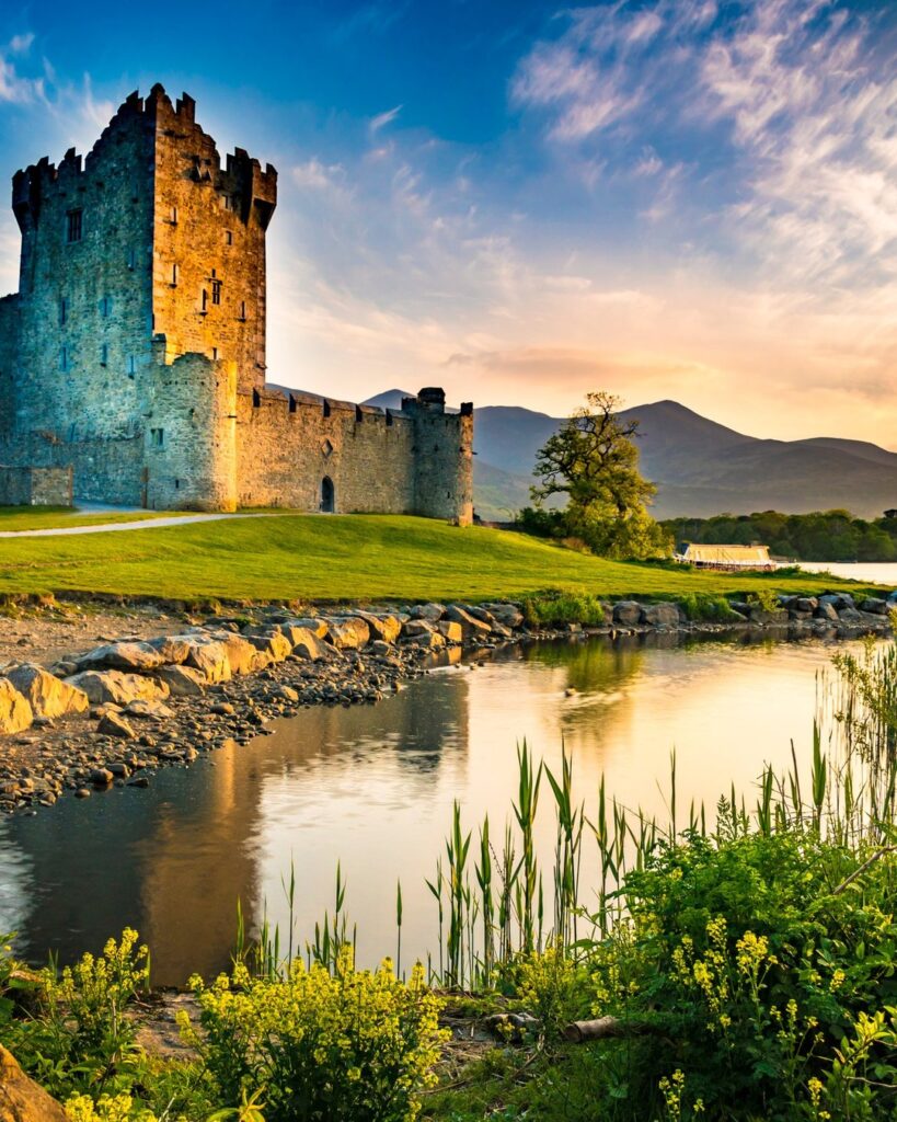 Medieval stone castle tower beside calm river at golden hour with green hills