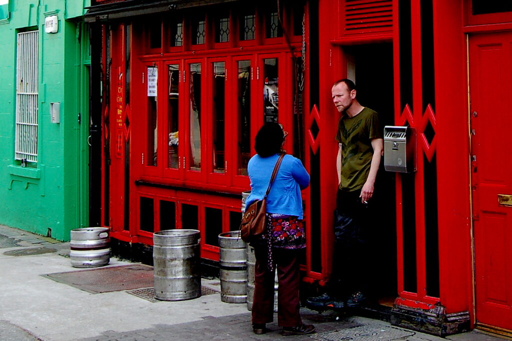 Two people conversing outside a red-painted building with metal bollards on the sidewalk