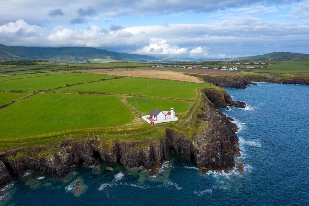 White lighthouse on green clifftop peninsula surrounded by blue ocean and rolling green fields