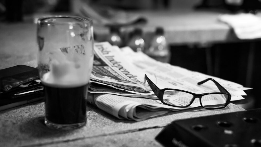 Black and white photo of a pint glass with dark beer beside folded newspapers and eyeglasses on a table