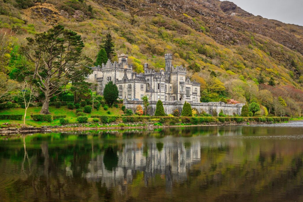 White castle with towers reflected in calm river, surrounded by green hillside forest