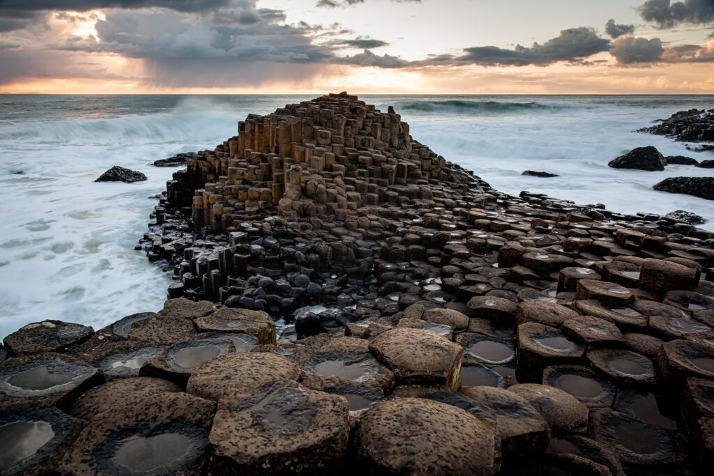 Hexagonal basalt columns extend into the sea under a dramatic cloudy sunset