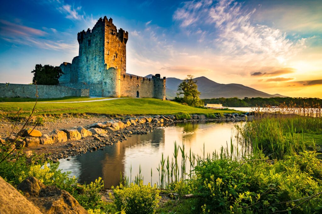 Medieval stone castle tower beside river at sunset with mountains in background
