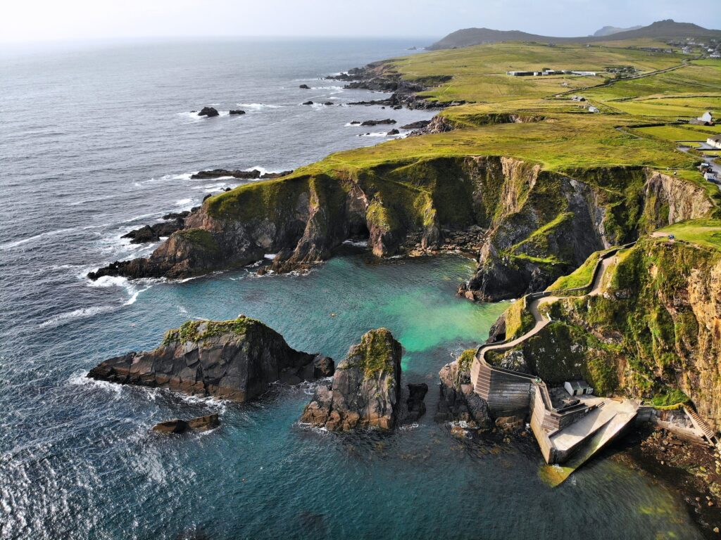 Turquoise cove beneath moss-covered sea cliffs with wooden bridge and green hillside