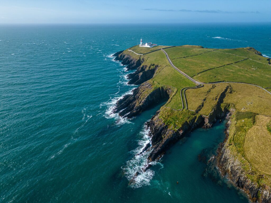 Aerial view of green coastal headland with lighthouse and winding road beside turquoise sea