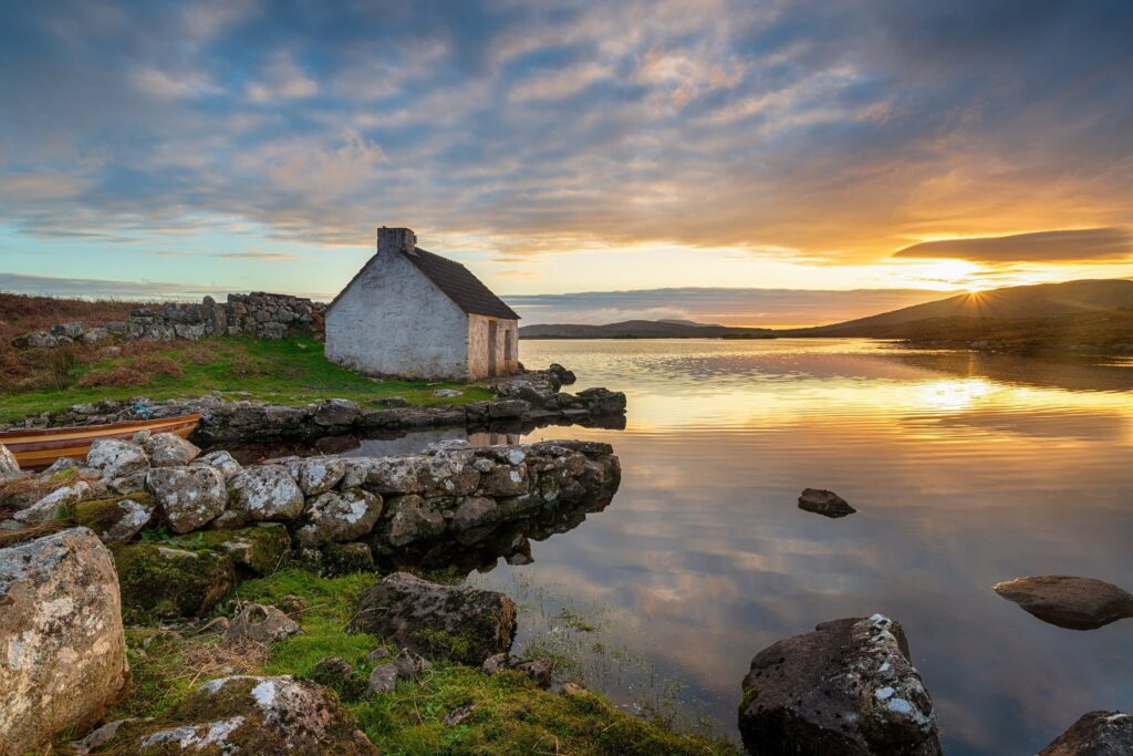 White cottage on rocky shoreline at sunset with golden light reflecting on calm water