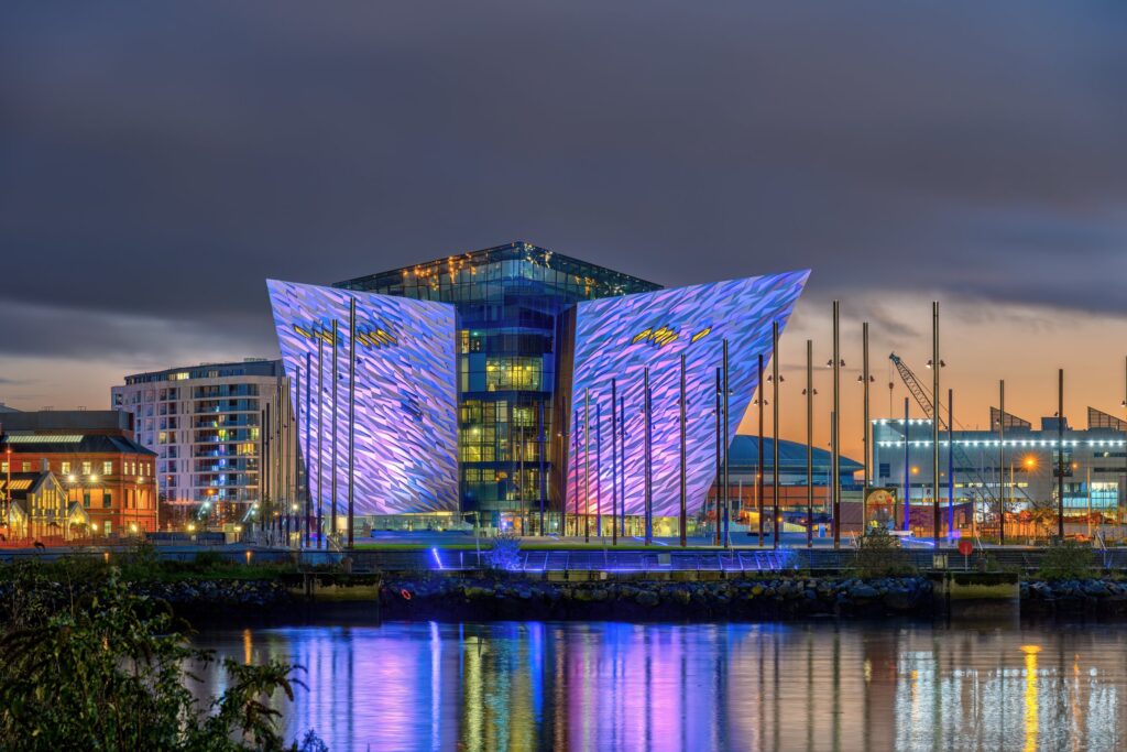 Illuminated modern waterfront building with blue and purple facade reflecting in still water at dusk