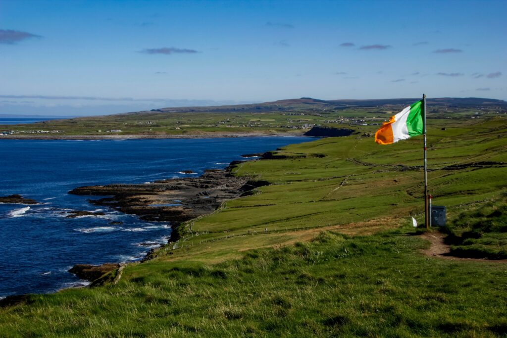 Irish flag flying on coastal headland overlooking green fields and rocky shore