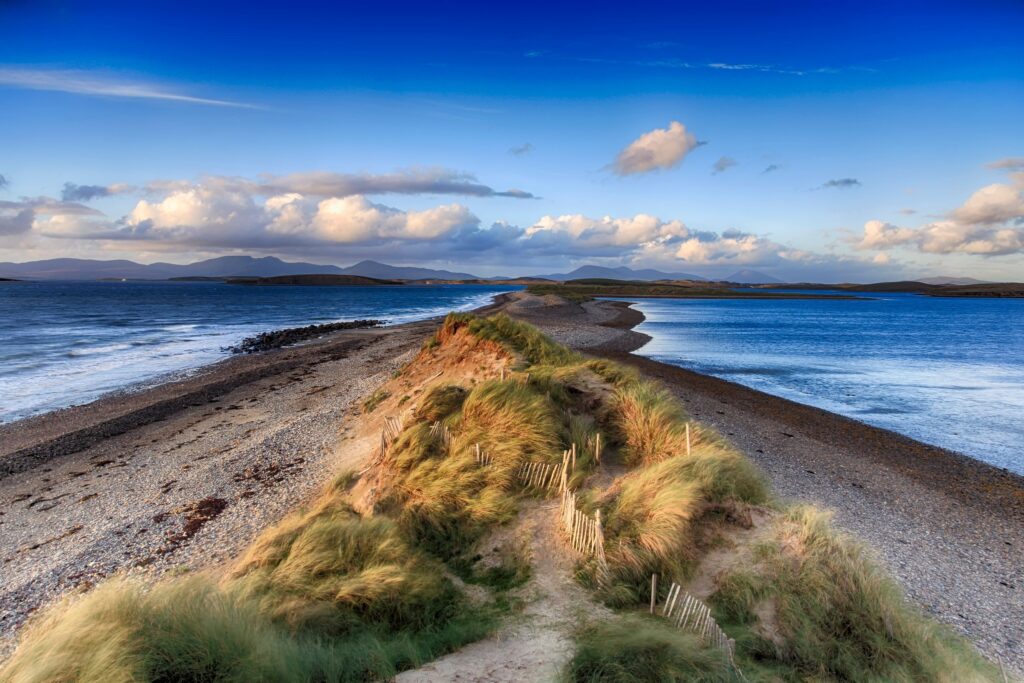 Grassy sand dune peninsula with wooden fencing overlooking blue ocean and distant mountains